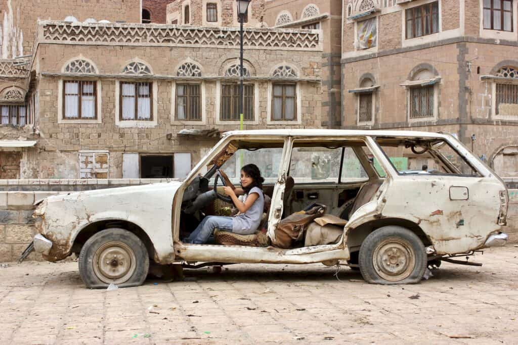 Photo of a girl sitting in a decrepit car in front of a building in Yemen